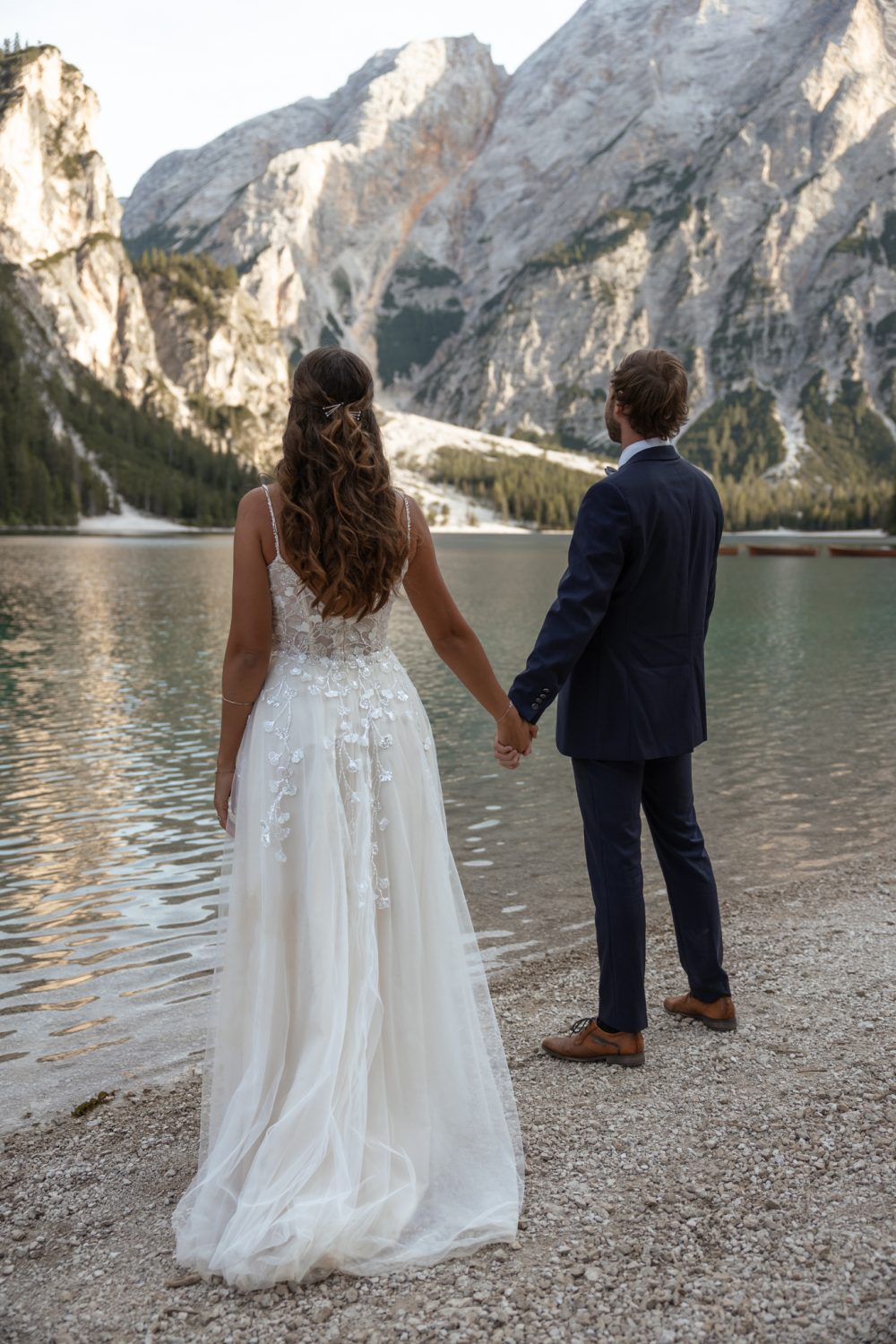 Braut und Bräutigam stehen Hand in Hand am Ufer des Pragser Wildsees und schauen auf die Berge