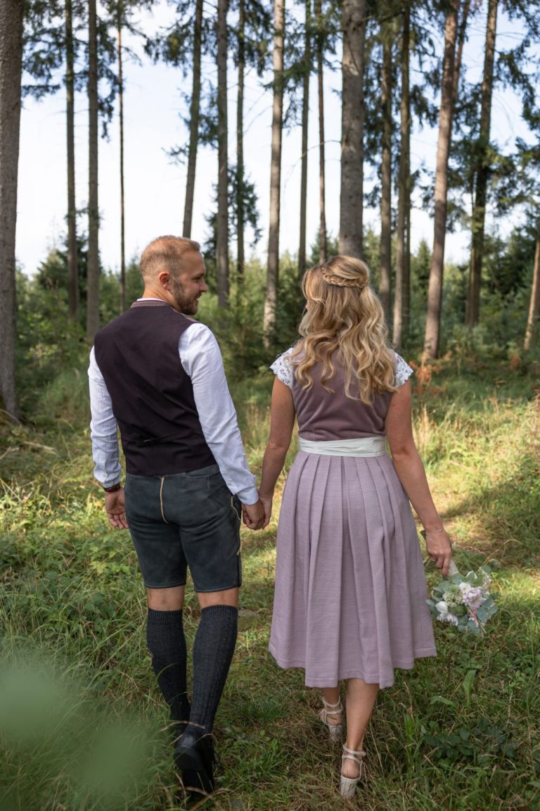 Brautpaar in Tracht geht Hand in Hand durch einen Wald, von hinten gesehen