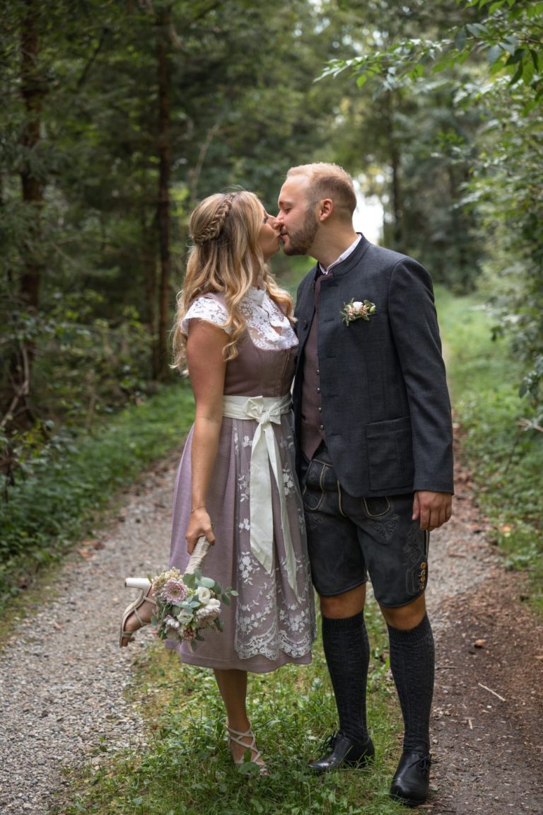 Brautpaar in Tracht küsst sich auf einem Waldweg, sie hält einen Brautstrauß