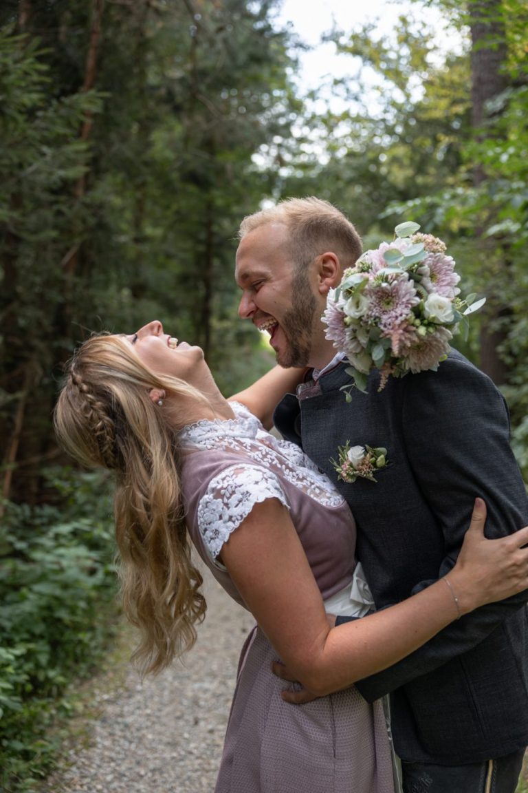 Brautpaar in Tracht umarmt sich lachend im Wald, er hält den Brautstrauß