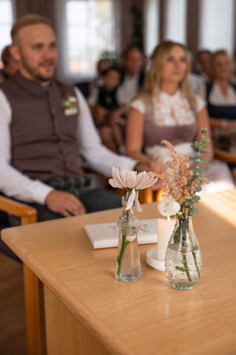 Trautisch mit Blumenvasen und Kerze, dahinter das Brautpaar in Tracht und Gäste