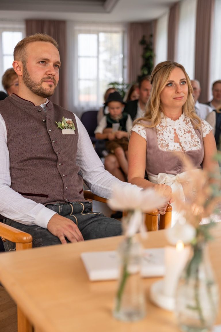 Brautpaar in Tracht sitzt während der Trauung am Tisch, dahinter Gäste und große Fenster