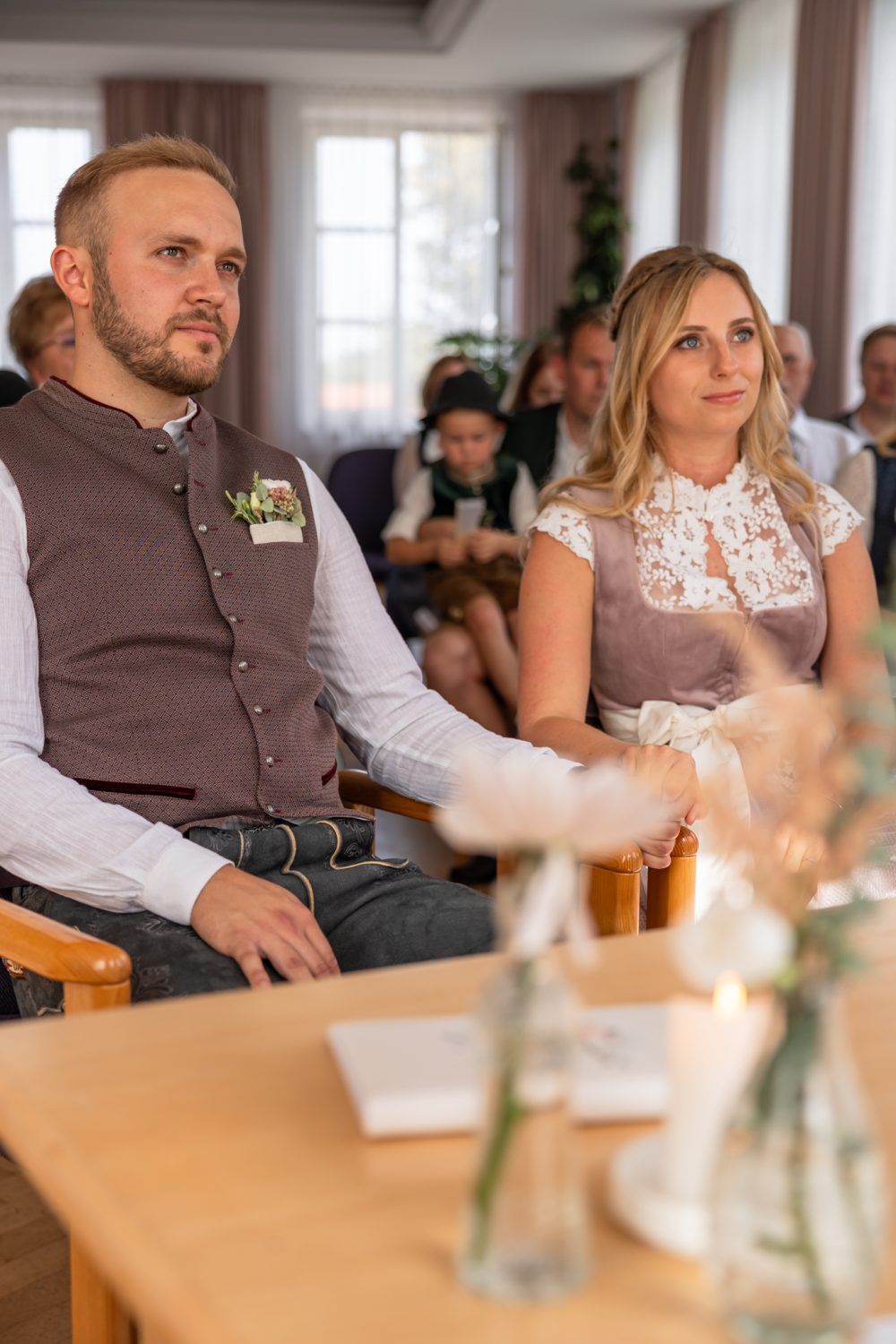 Brautpaar in Tracht sitzt während der Trauung am Tisch, dahinter Gäste und große Fenster