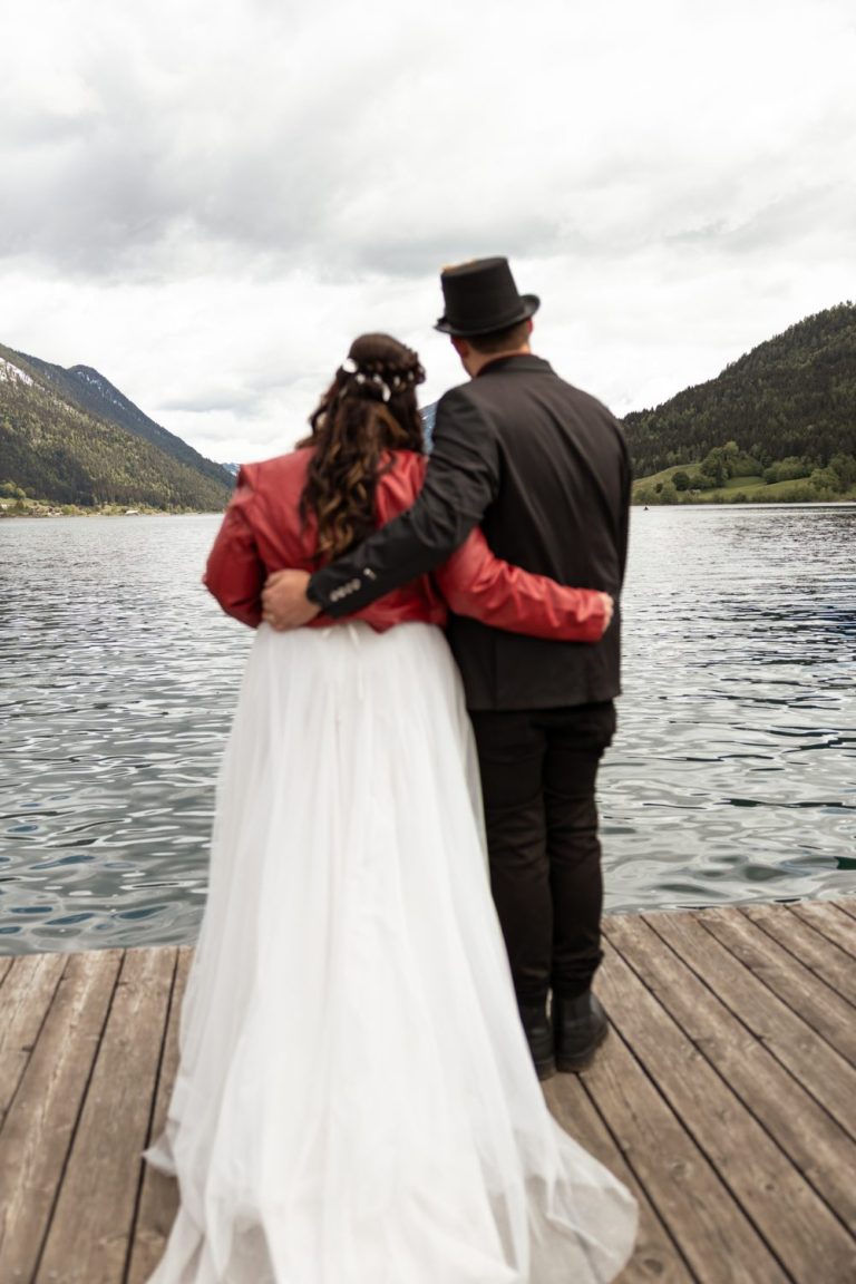 Brautpaar von hinten auf einem Holzsteg am See, sie in rotem Jäckchen, er mit Hut, dahinter Berge