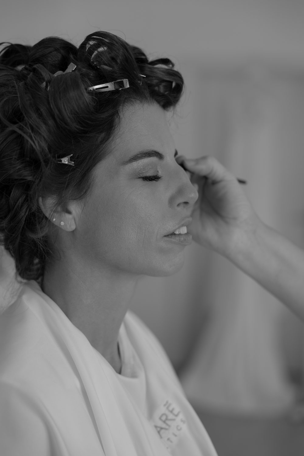 Braut mit Lockenwicklern im Haar tupft sich beim Make-up an die Augenbraue (Schwarz-Weiß-Aufnahme)