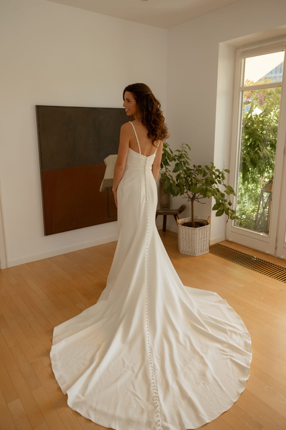Braut im Hochzeitskleid steht von hinten in einem hellen Zimmer neben einem großen Fenster