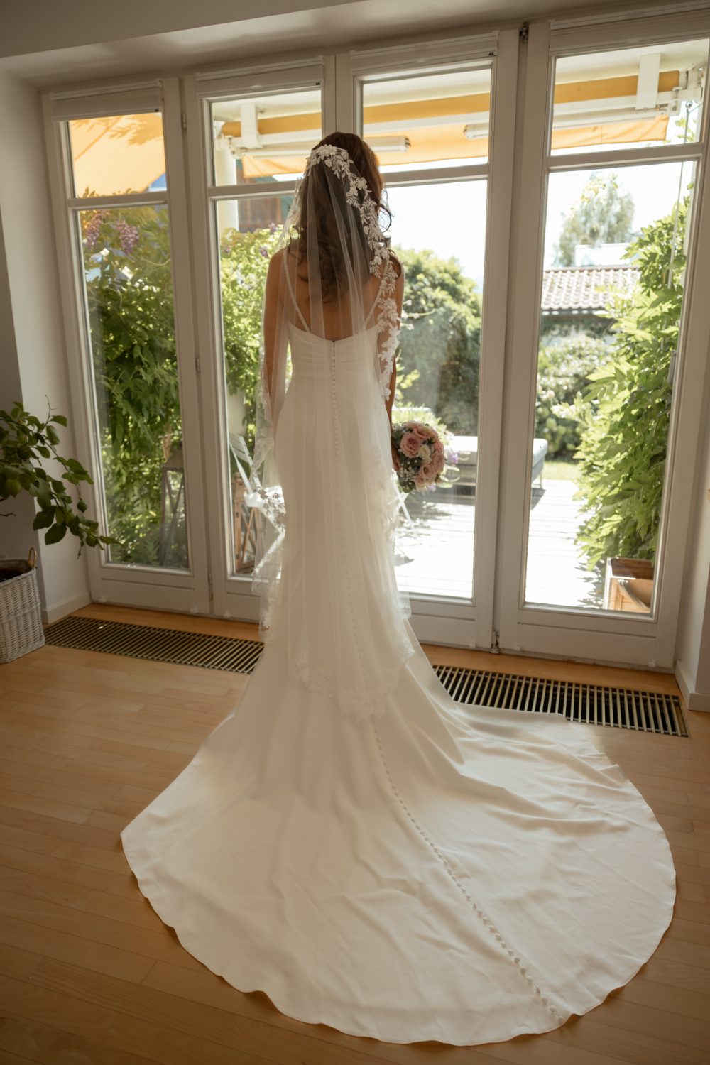 Braut im Hochzeitskleid mit langer Schleppe steht von hinten vor einer bodentiefen Fenstertür