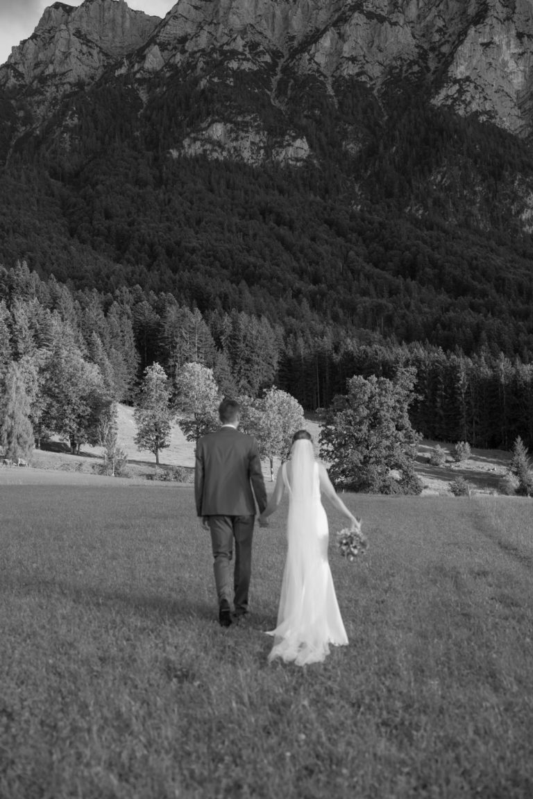 Brautpaar geht Hand in Hand über eine Wiese, dahinter Wald und Felswand (Schwarz-Weiß-Aufnahme)