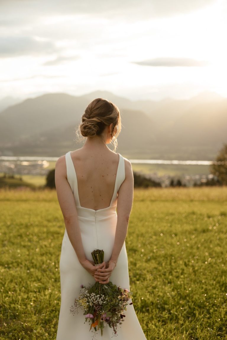 Braut im rückenfreien Kleid von hinten auf einer Wiese, den Brautstrauß hinter dem Rücken, dahinter Berge