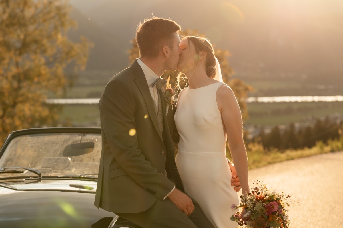 Franzi und Christoph küssen sich im Abendlicht neben einem Cabrio