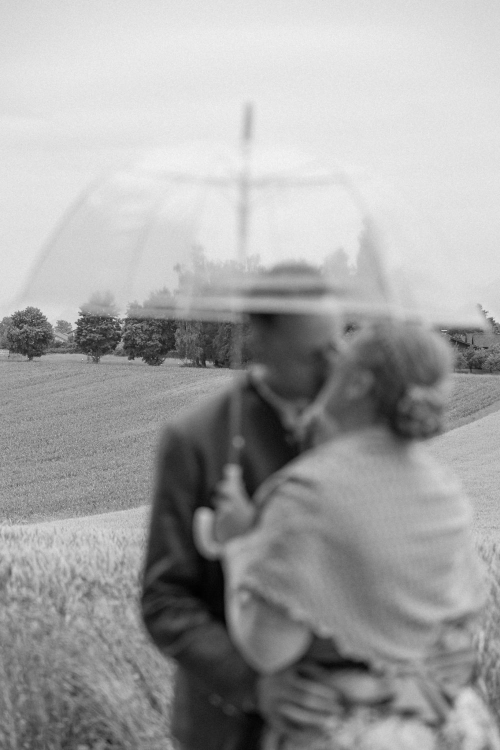Paar umarmt sich unter einem durchsichtigen Regenschirm vor einem Feld (Schwarz-Weiß-Aufnahme)