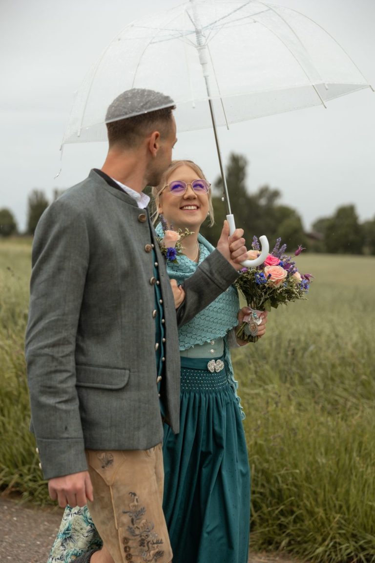 Bräutigam mit Regenschirm und lächelnde Braut in Tracht mit Brautstrauß vor einem Feld