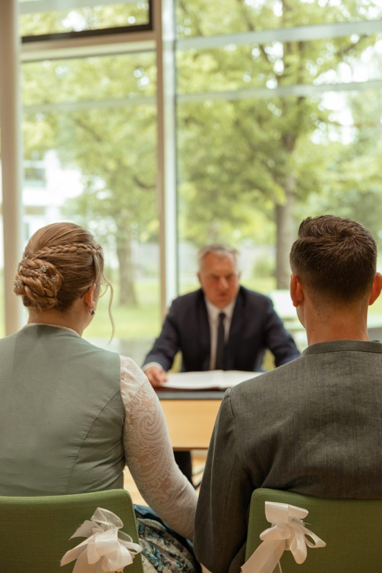 Brautpaar sitzt von hinten vor dem Standesbeamten an einem Tisch im Trauzimmer, dahinter große Fenster