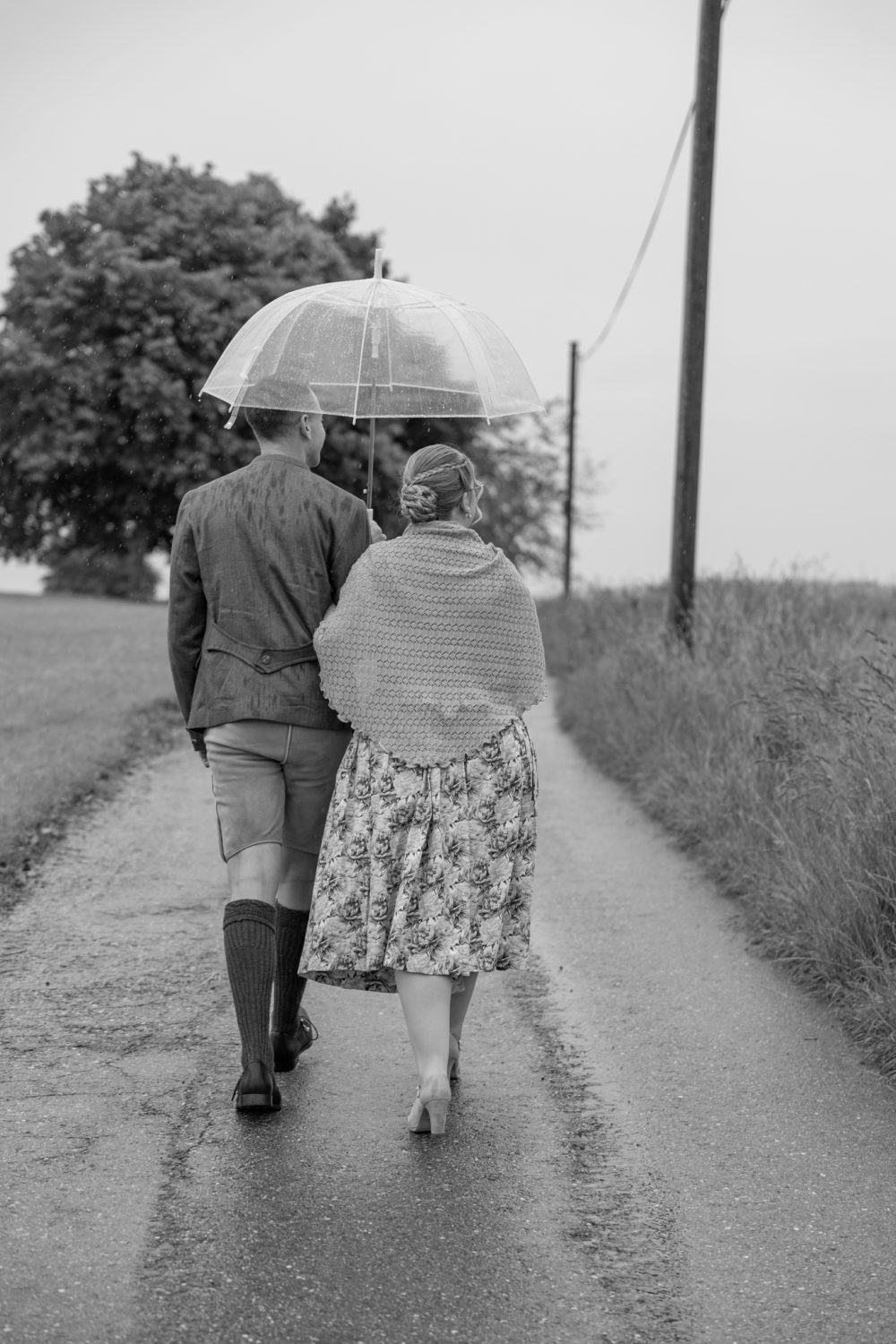 Paar geht unter einem durchsichtigen Regenschirm eine nasse Straße entlang, von hinten gesehen (Schwarz-Weiß-Aufnahme)