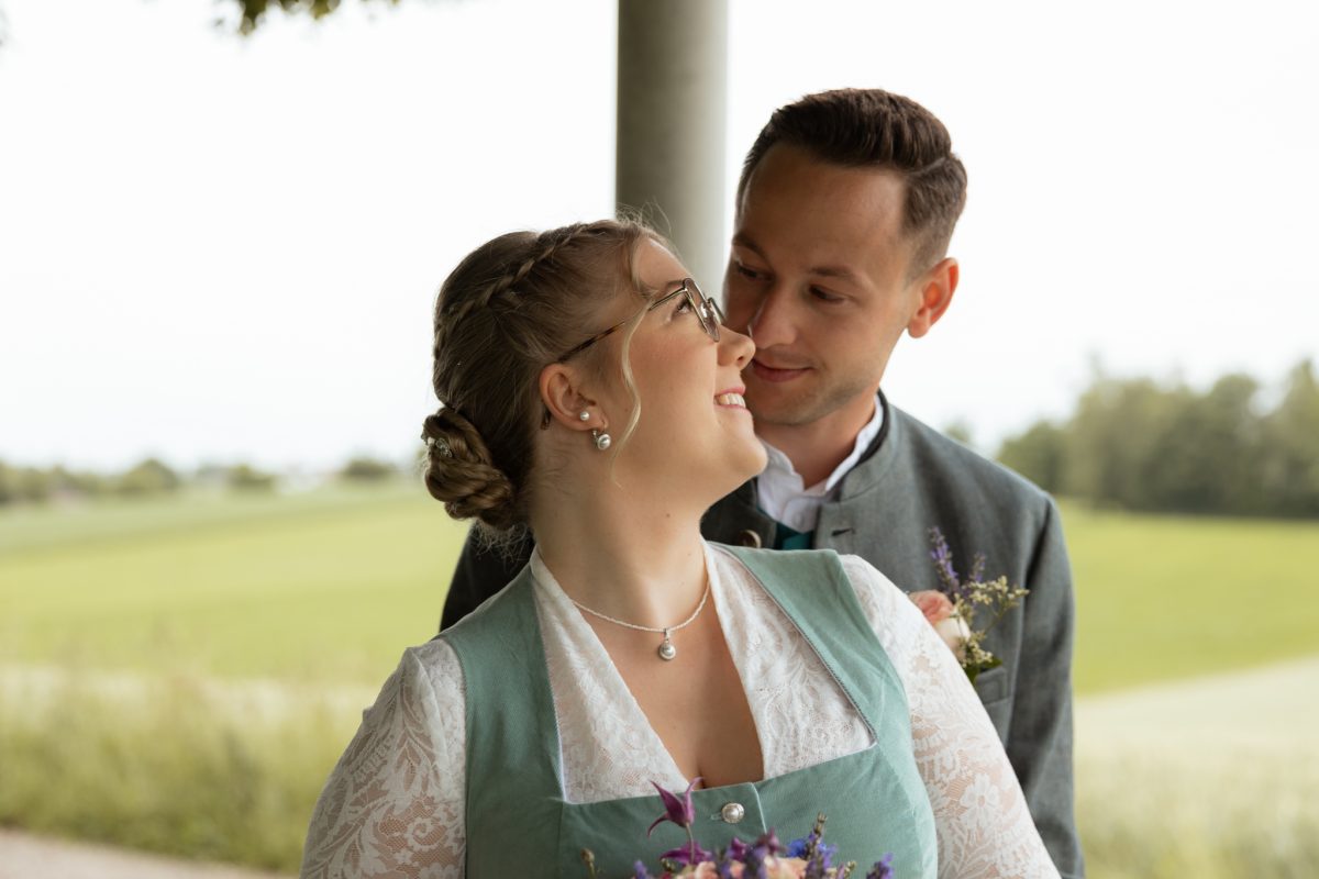 Braut in Tracht blickt lächelnd zum Bräutigam, dahinter Felder
