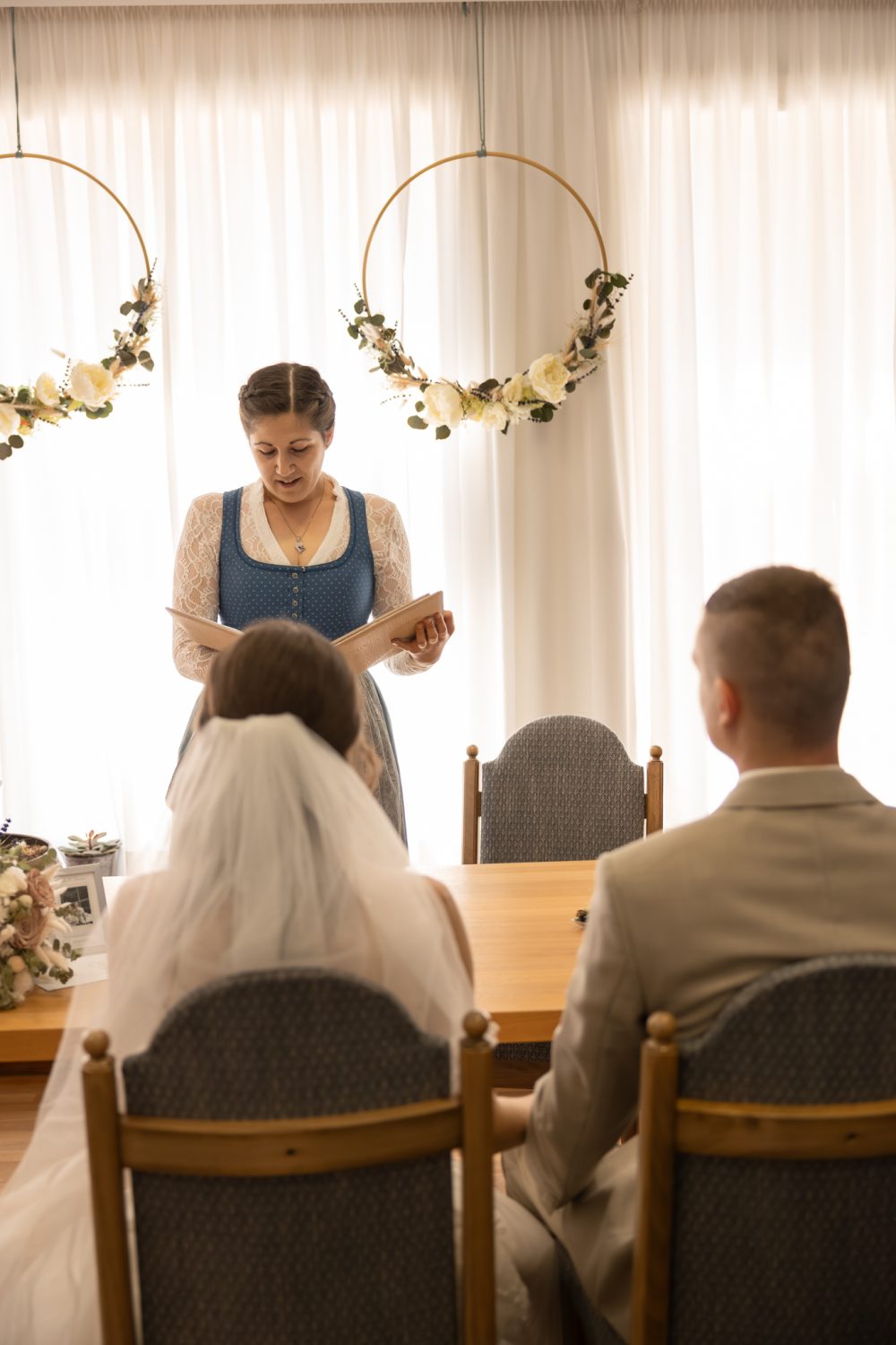 Frau im Dirndl liest vor dem Brautpaar am Tisch aus einem Heft vor, dahinter zwei Blumenkränze am Vorhang