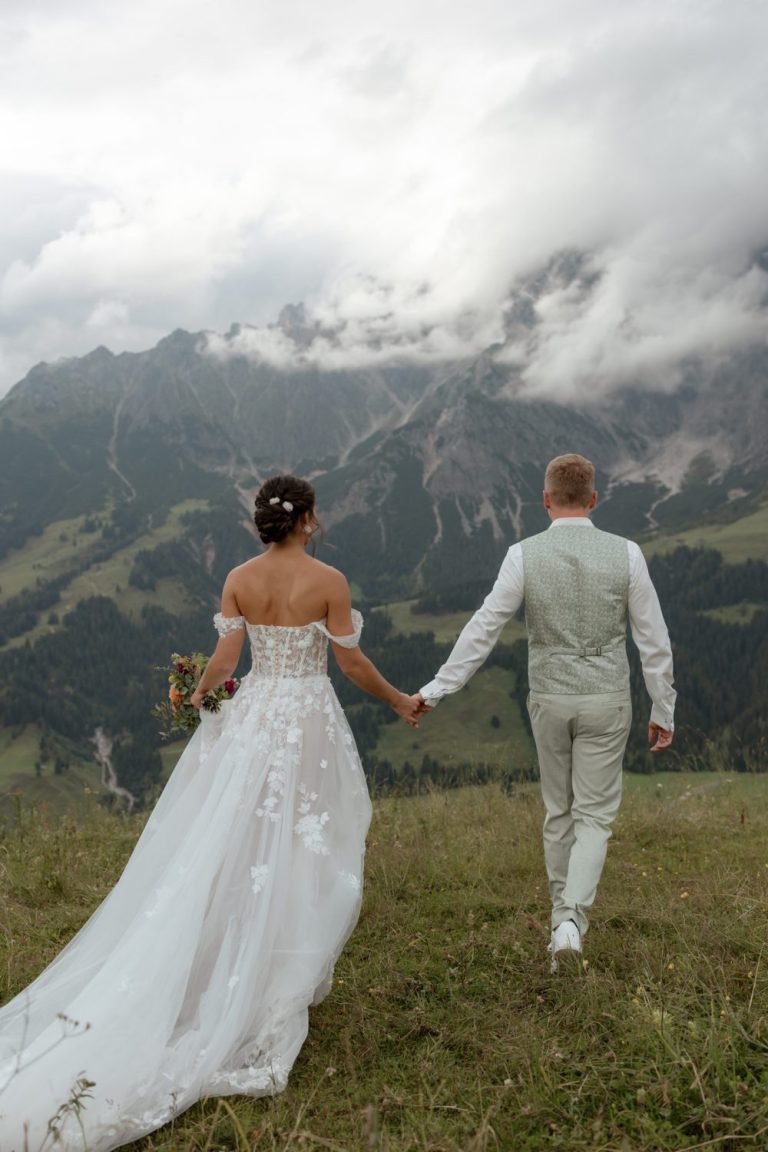 Brautpaar geht Hand in Hand über eine Bergwiese auf Berge zu, von hinten gesehen