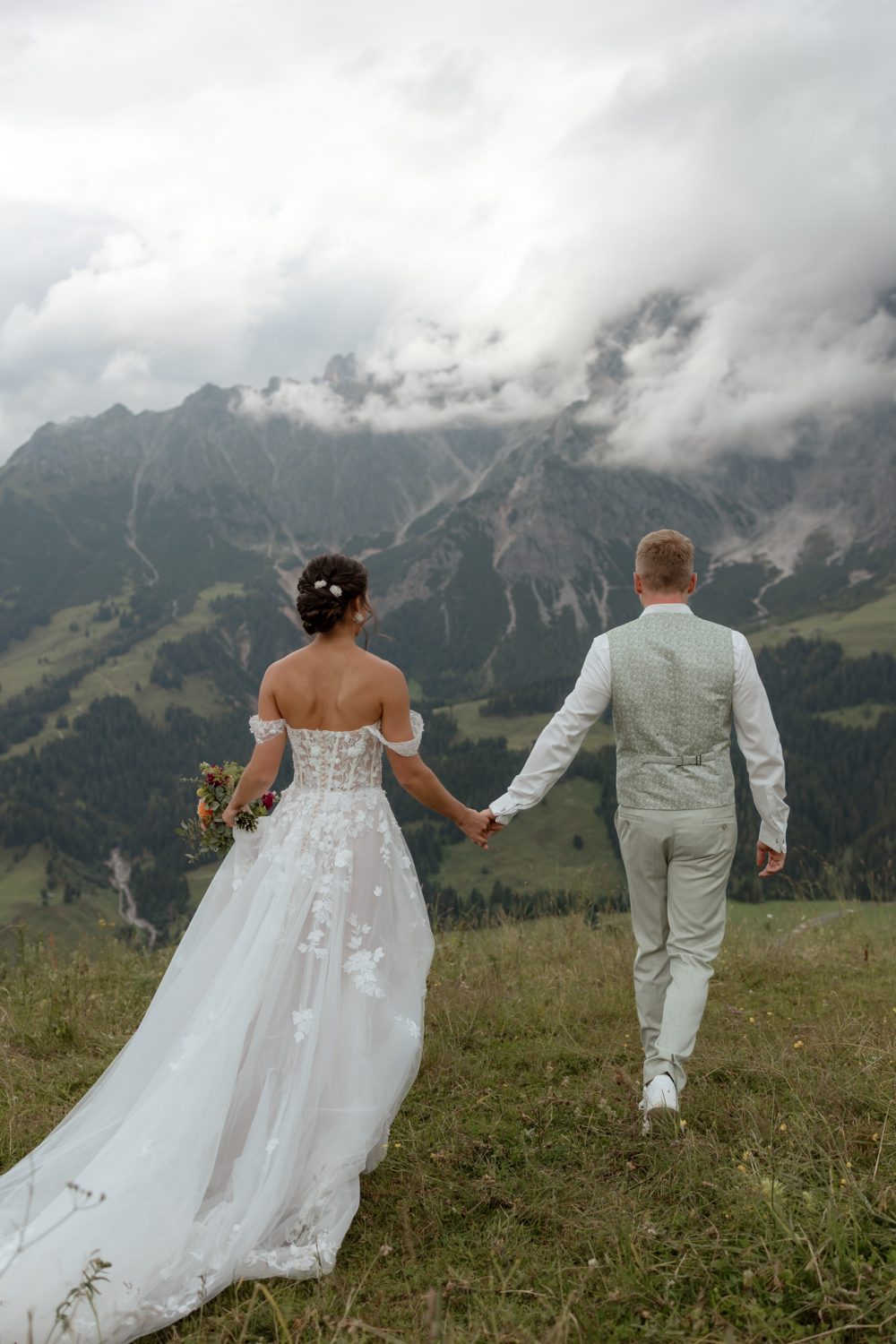 Brautpaar geht Hand in Hand über eine Bergwiese auf Berge zu, von hinten gesehen