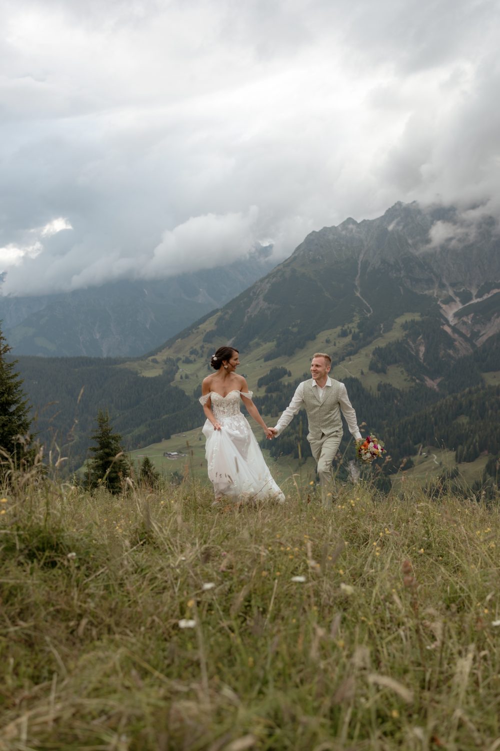 Brautpaar geht Hand in Hand einen Berghang hinunter, im Hintergrund Berge und Wolken