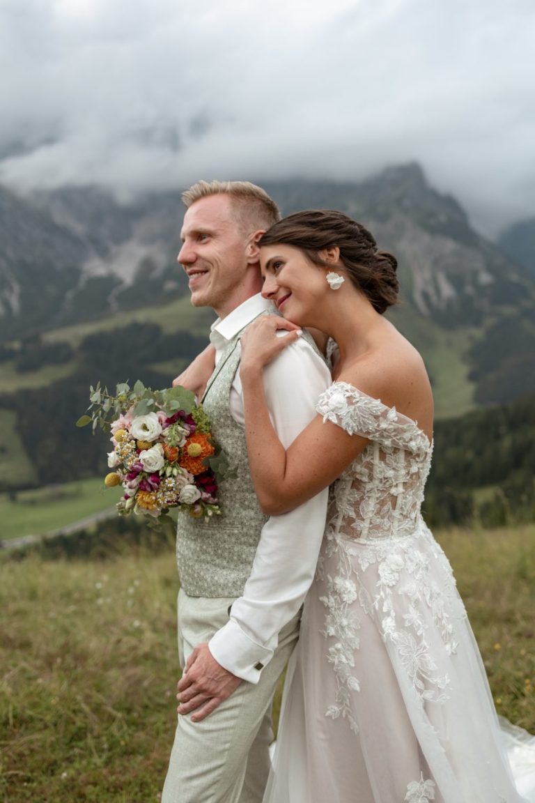 Braut lehnt sich an den Bräutigam und hält ihren Brautstrauß, dahinter Berge