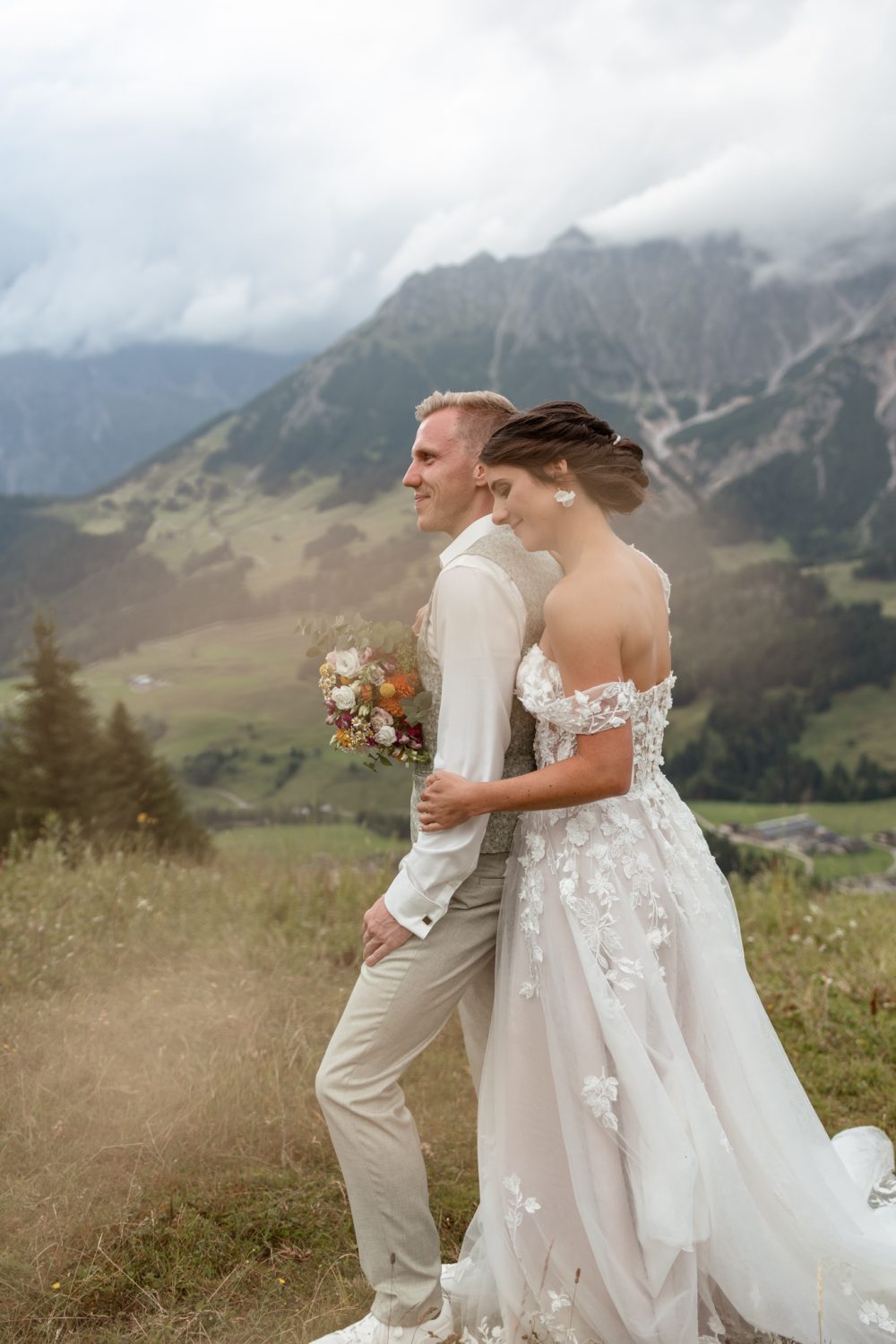 Brautpaar im Gras vor Bergpanorama, er im hellen Anzug, sie im Spitzenkleid