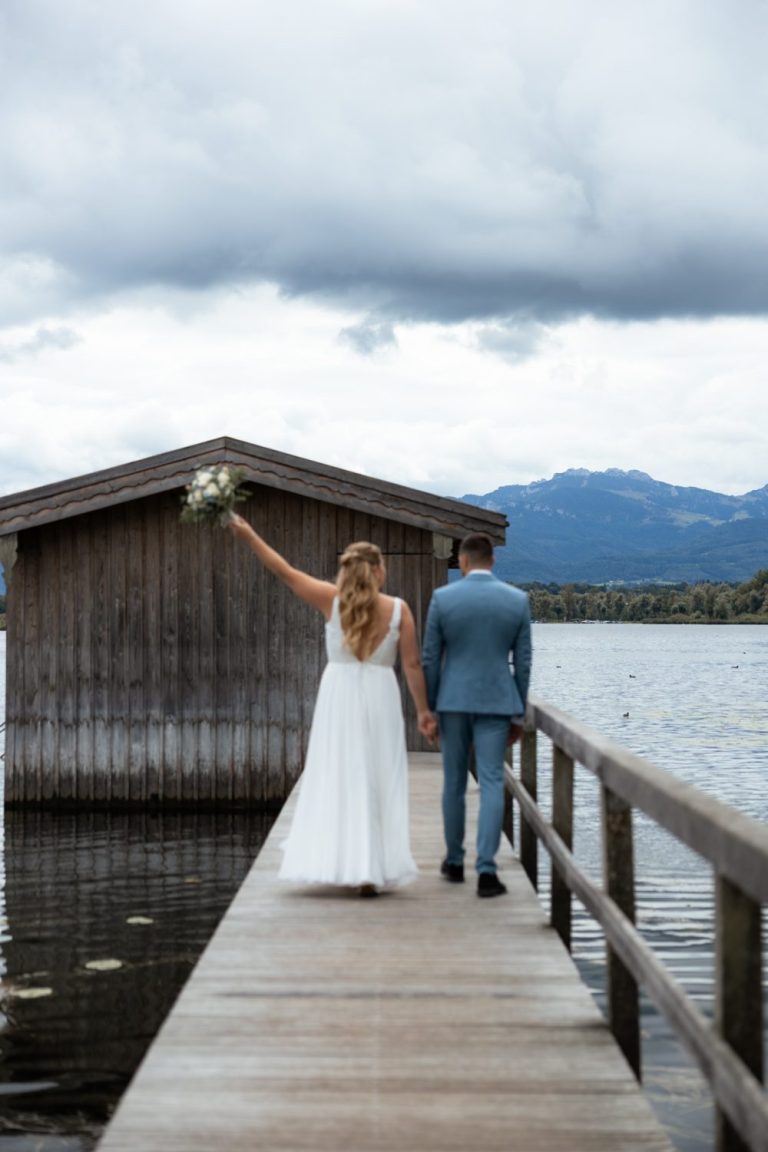 Braut streckt den Brautstrauß in die Höhe, beide gehen von hinten über einen Holzsteg am See, dahinter eine Holzhütte und Berge