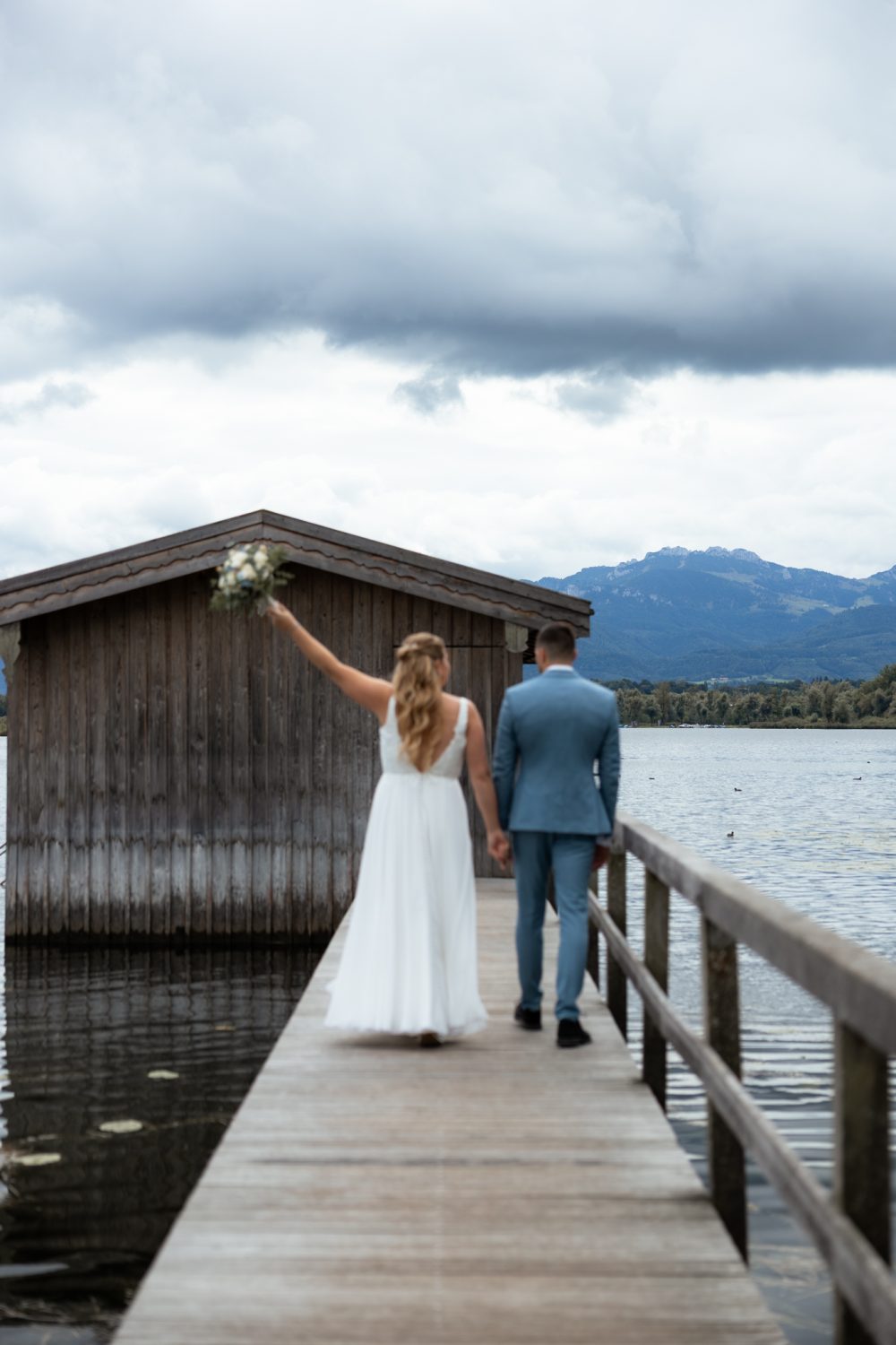Braut streckt den Brautstrauß in die Höhe, beide gehen von hinten über einen Holzsteg am See, dahinter eine Holzhütte und Berge