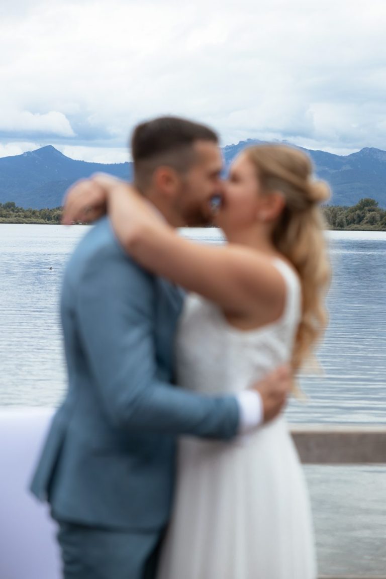 Brautpaar küsst sich am See, er im blauen Anzug, im Hintergrund Berge