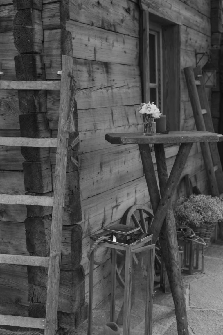 Rustikale Holzhütte mit Leiter, Stehtisch und kleiner Blumenvase (Schwarz-Weiß-Aufnahme)