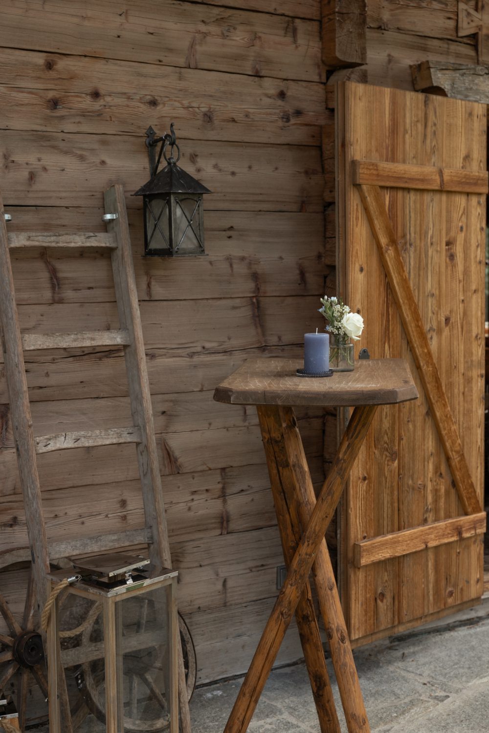 Holzwand mit Leiter, Laterne, Holztür und Stehtisch mit kleiner Blumenvase