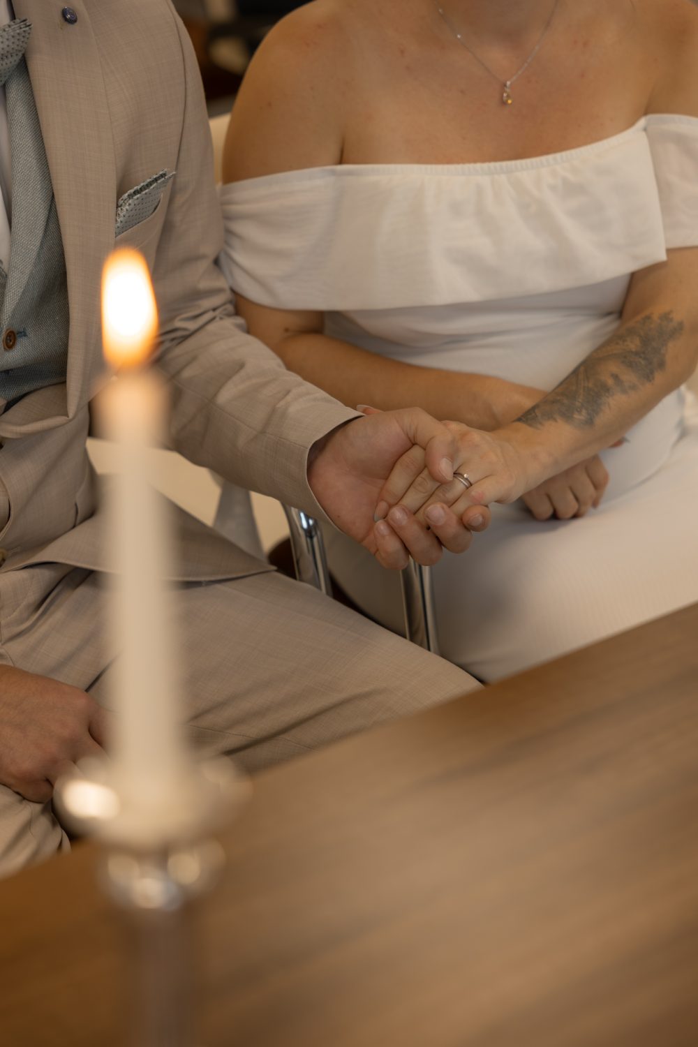 Ineinandergelegte Hände von Braut und Bräutigam am Tisch, davor eine brennende Kerze