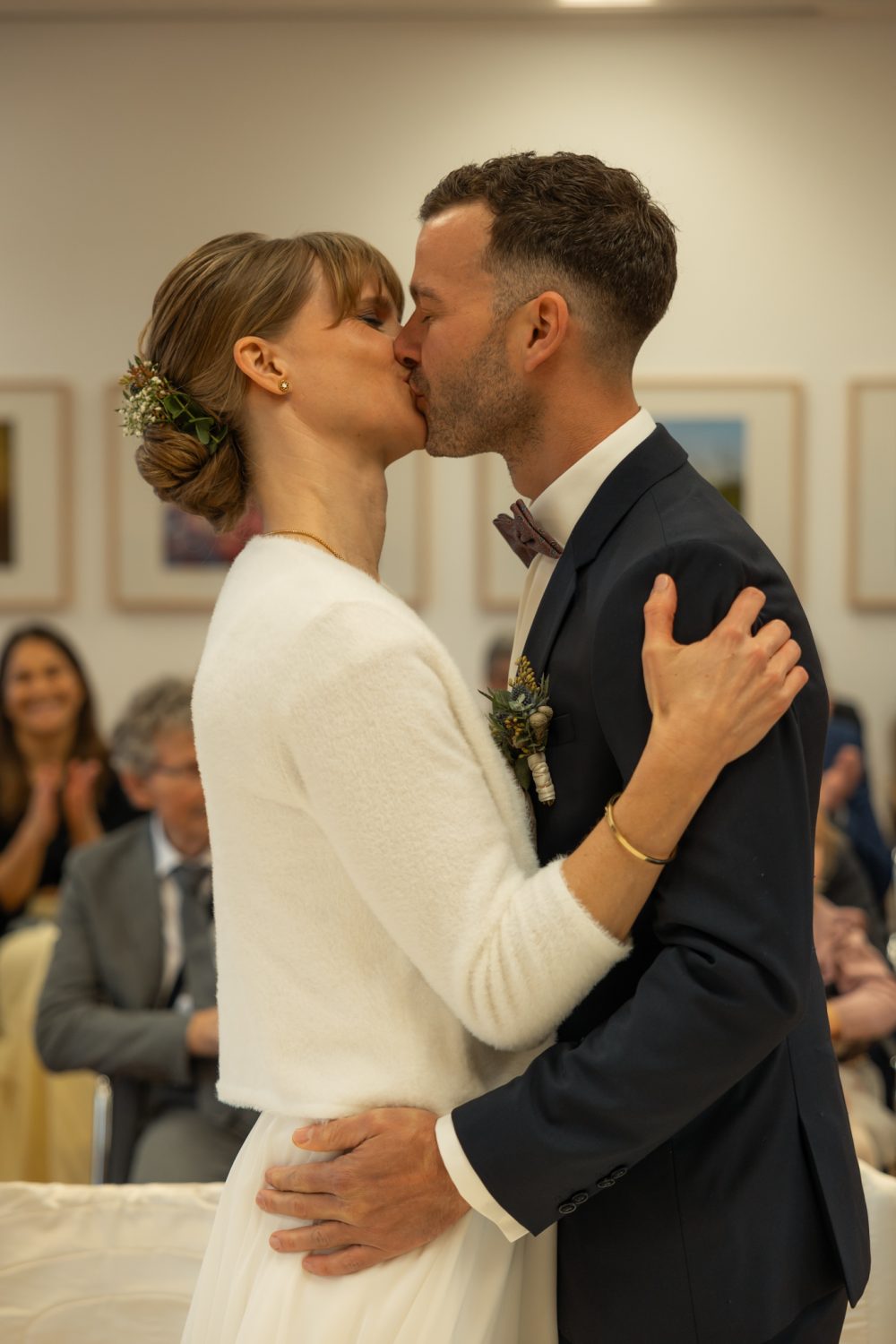 Brautpaar küsst sich, sie mit Blumenhaarschmuck und heller Jacke, er im dunklen Anzug mit Fliege, im Hintergrund Gäste