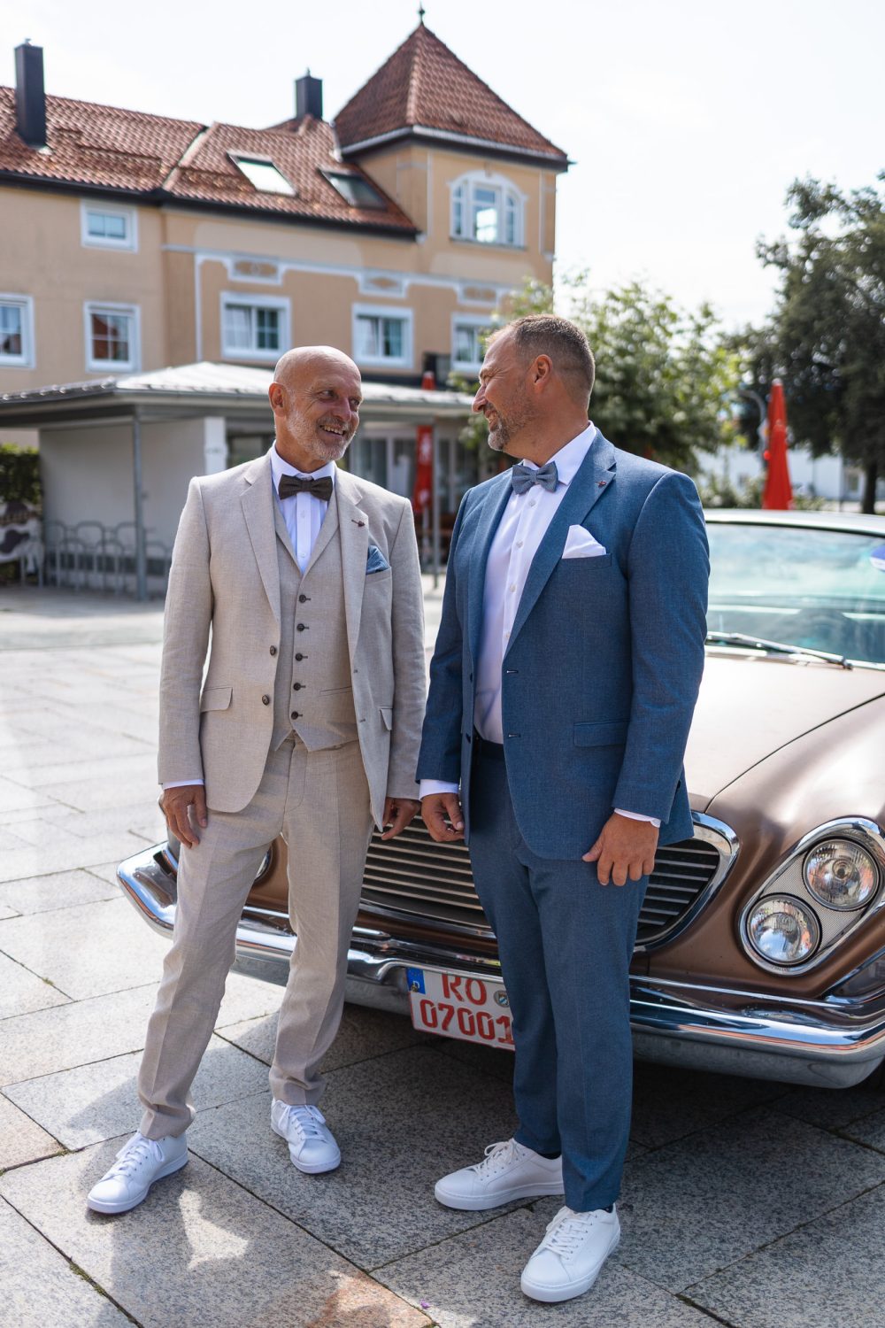 Zwei Männer in Anzug und Fliege stehen lachend vor einem Oldtimer auf einem Platz