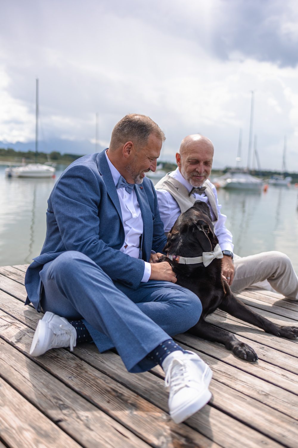 Zwei Männer im Anzug sitzen mit einem schwarzen Hund mit Fliege auf einem Holzsteg am Wasser, im Hintergrund Segelboote