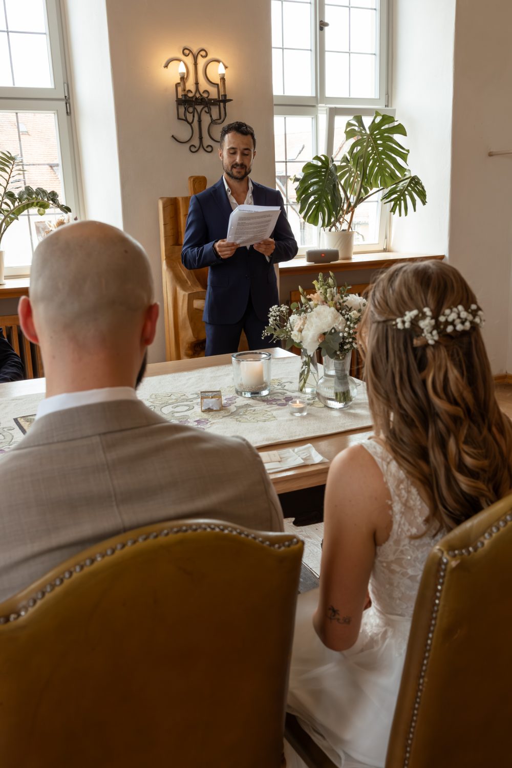 Rede im Trauzimmer vor dem sitzenden Brautpaar, auf dem Tisch Blumenstrauß und Kerzen