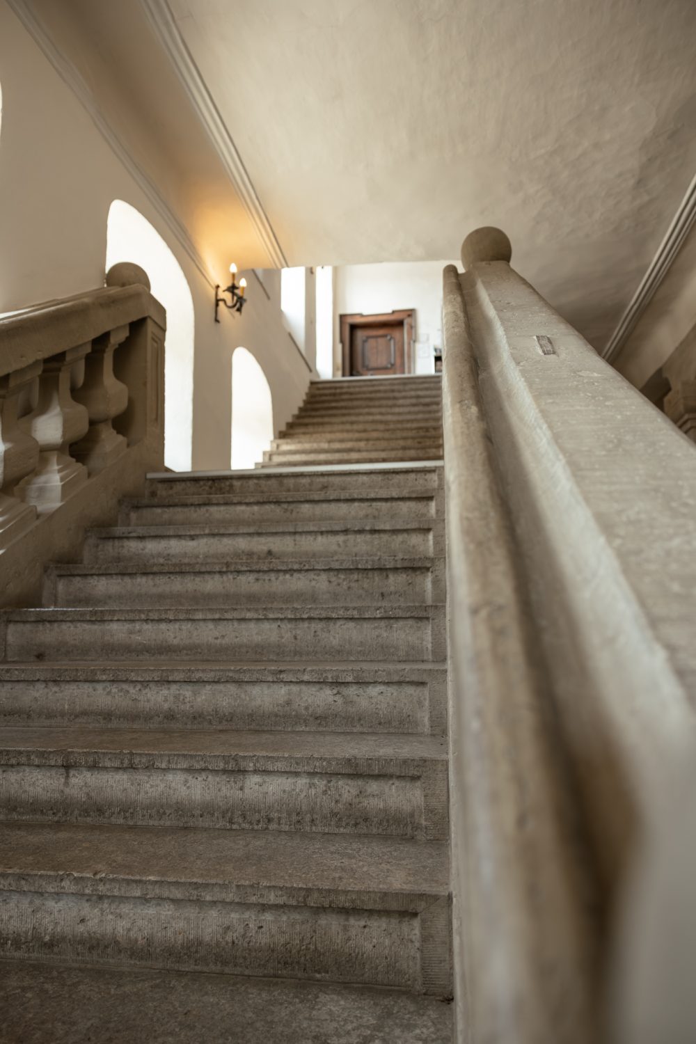 Steintreppe mit Geländer im hellen Treppenhaus, oben eine Holztür